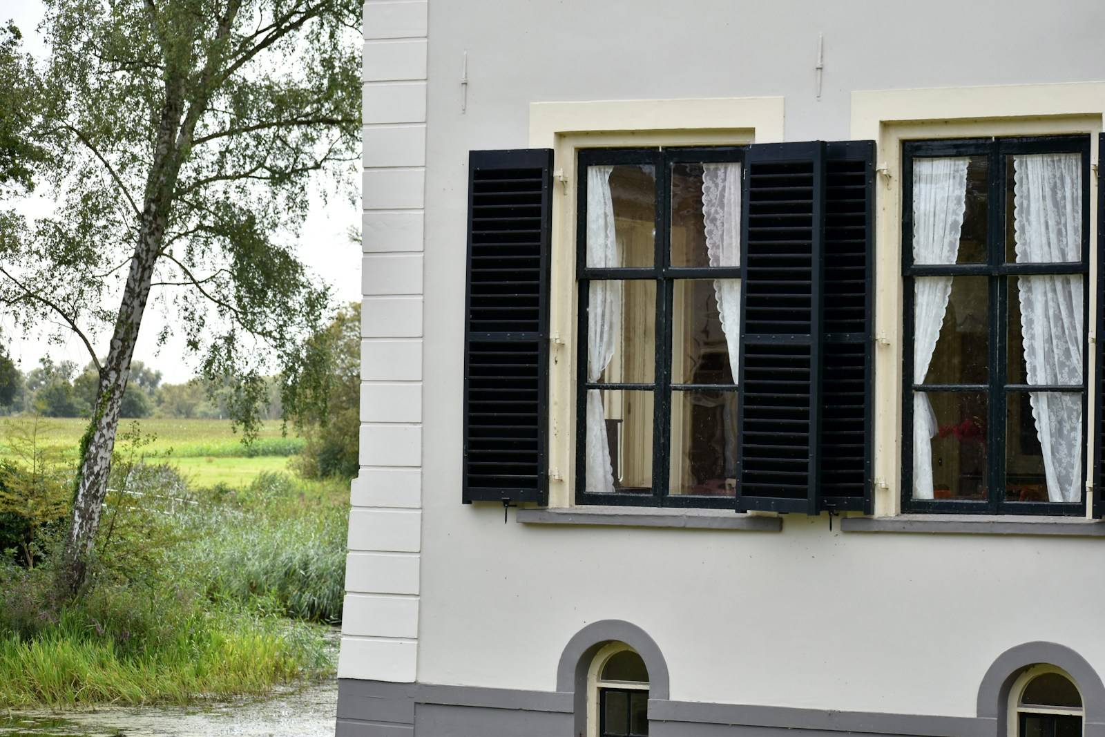 Witte gepleisterde gevel met zwarte luiken, met bomen ernaast
